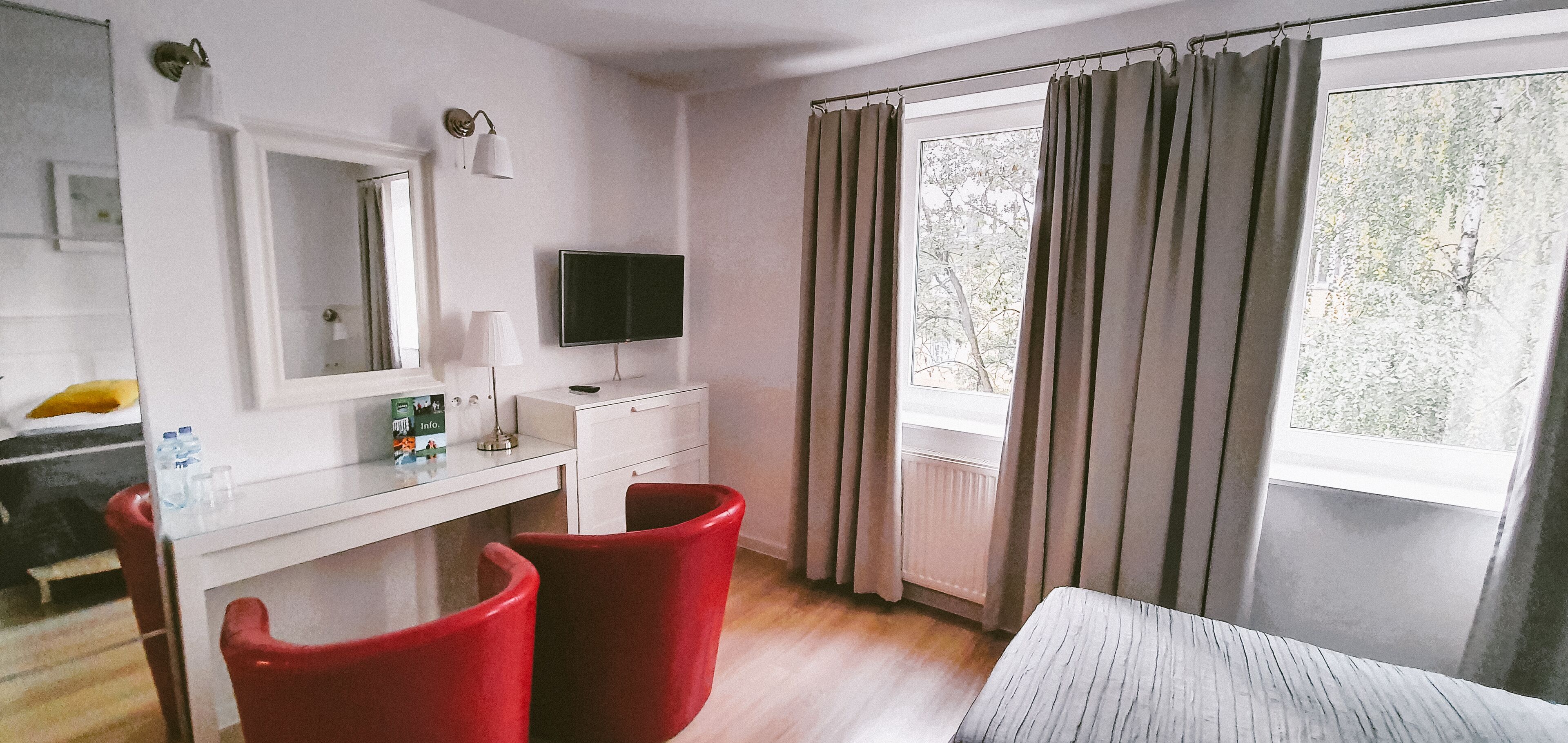 A hotel room with a white desk, two red chairs, a flat-screen TV, a mirror, and a window with curtains.