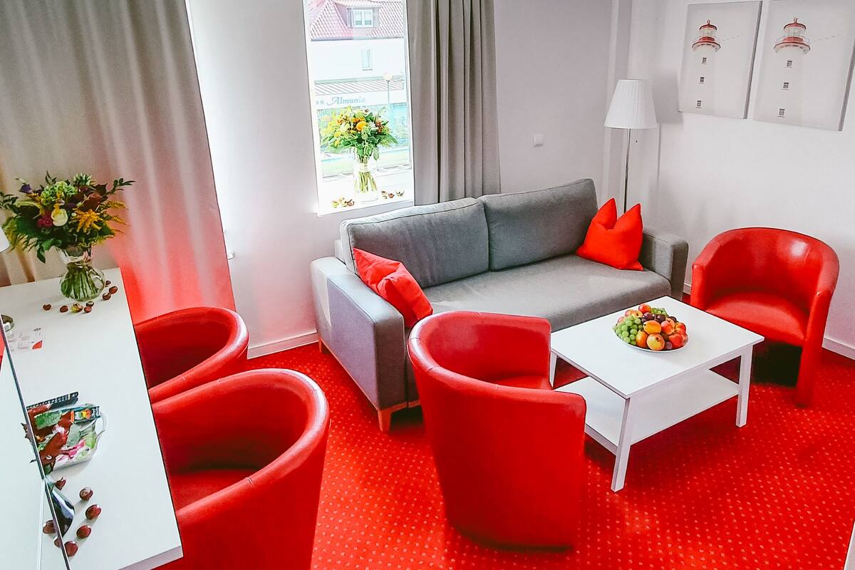 A modern living room with a grey sofa, red chairs, a white coffee table, and a window with a view of a building.
