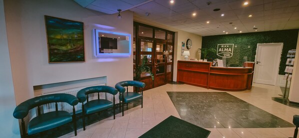 Zona de recepción con mostrador, un reloj y una pared verde con un cartel. Hay tres sillas azules y un mostrador de recepción.