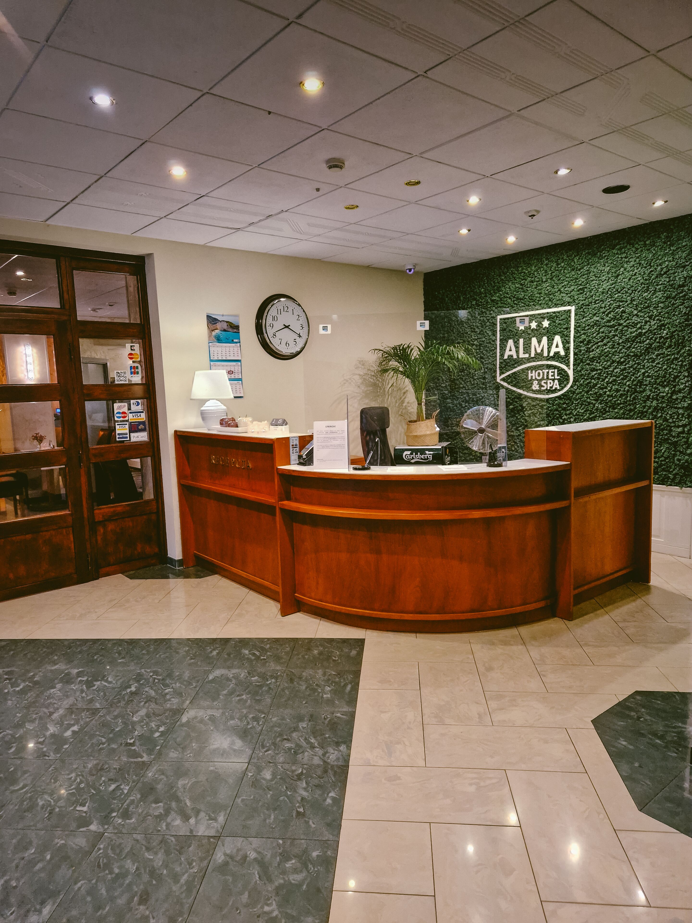 Un comptoir d’accueil avec une horloge, le logo de l’hôtel ALMA HOTEL & SPA et un mur végétal.