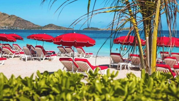 A beach with red umbrellas and white lounge chairs, a clear blue sea, and a mountain in the background.