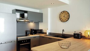 A modern kitchen with a stainless steel refrigerator, built-in oven, microwave, and coffee maker. There's a wooden countertop, a sink, and a large wall clock.