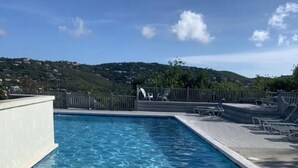 A clear blue pool with a wooden deck and lounge chairs, surrounded by a white wall and a view of a green hillside.