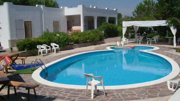 Una piscina con zona ajardinada blanca, sillas de descanso y una zona de estar cubierta.