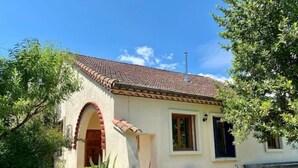 Ein einstöckiges Haus mit einem Ziegeldach, einer kleinen Veranda mit zwei Stühlen und einem Gartenbereich mit verschiedenen Pflanzen.