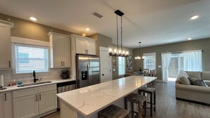 A modern kitchen with white cabinets, a stainless steel refrigerator, a marble island, wooden stools, and a dining area with a sofa and curtains. - CHIll IN! Central to Everything! Steps to Beach! (Corolla)