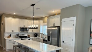 A modern kitchen with white cabinetry, a large island with bar stools, stainless steel appliances, and a pendant light fixture. - CHIll IN! Central to Everything! Steps to Beach! (Corolla)