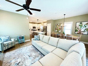 Un salon moderne avec un canapé d’angle, un ventilateur de plafond et une cuisine en arrière-plan.