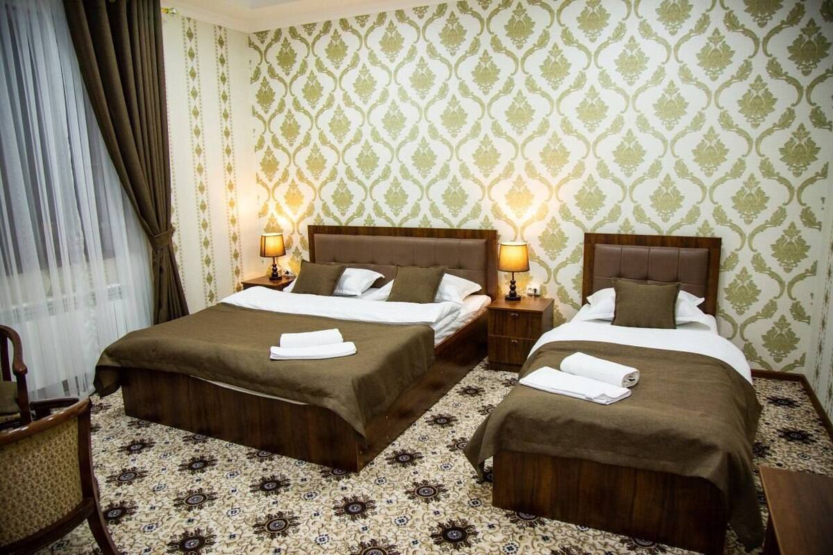 A hotel room with two beds, each with a wooden headboard, white bedding, and a brown throw blanket. There are two bedside lamps, a chair, and a patterned carpet.