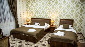 A hotel room with two beds, each with a wooden headboard, white bedding, and a brown throw blanket. There are two bedside lamps, a chair, and a patterned carpet.