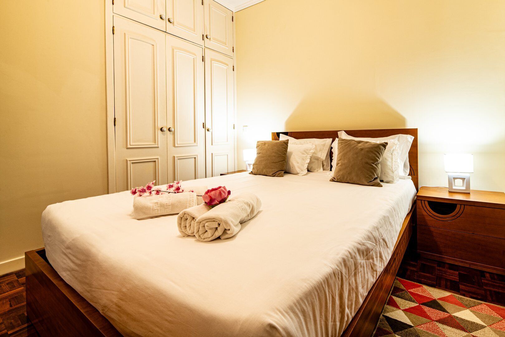 Una cama bien hecha con sábanas blancas y flores rosadas, un cabecero de madera y una mesita de noche con una lámpara.