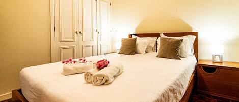 Una cama bien hecha con sábanas blancas y flores rosadas, un cabecero de madera y una mesita de noche con una lámpara.