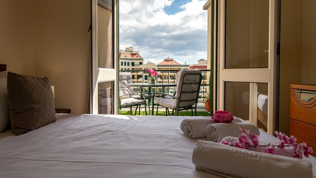 A hotel room with a bed, a chair, a table, and a balcony with a view of buildings. - GuestReady - Family-Friendly Haven in Funchal (Funchal)