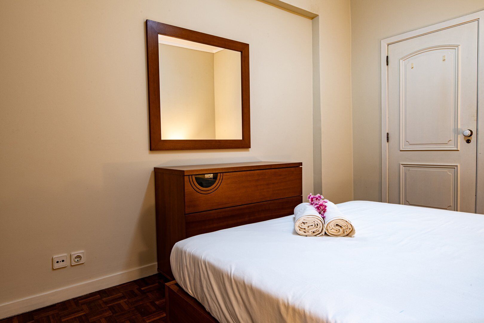Habitación de hotel con tocador de madera, cama con ropa blanca y espejo sobre el tocador, además de una puerta blanca.