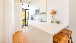 A modern kitchen with white cabinetry, a built-in oven, and a coffee machine. There is a small island with stools and a vase with dried plants.
