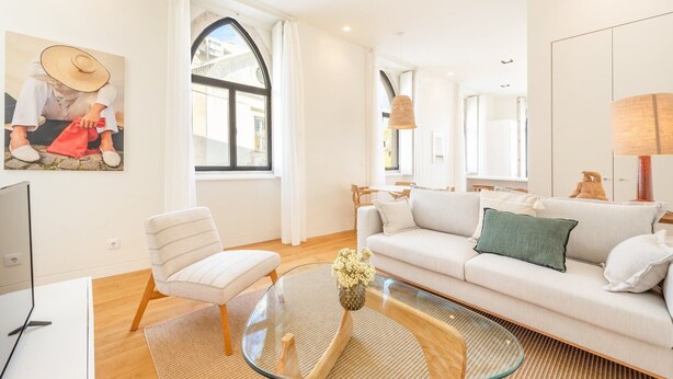 A modern living room with a white sofa, a glass coffee table, and a white armchair. There is a painting on the wall and a window with curtains.