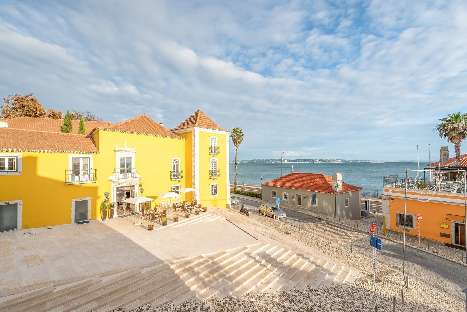 Un pueblo costero con edificios amarillos, un área pavimentada con mesas al aire libre y vistas al mar.