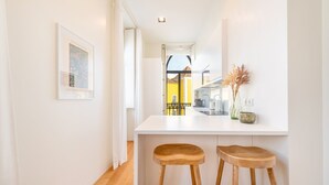 A narrow hallway with a white countertop and two wooden stools. A framed picture hangs on the wall, and a vase with dried plants is on the countertop.