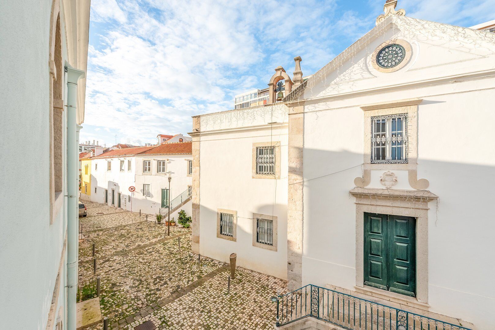 A narrow street with cobblestone pavement, white buildings with green doors, and a clear blue sky.