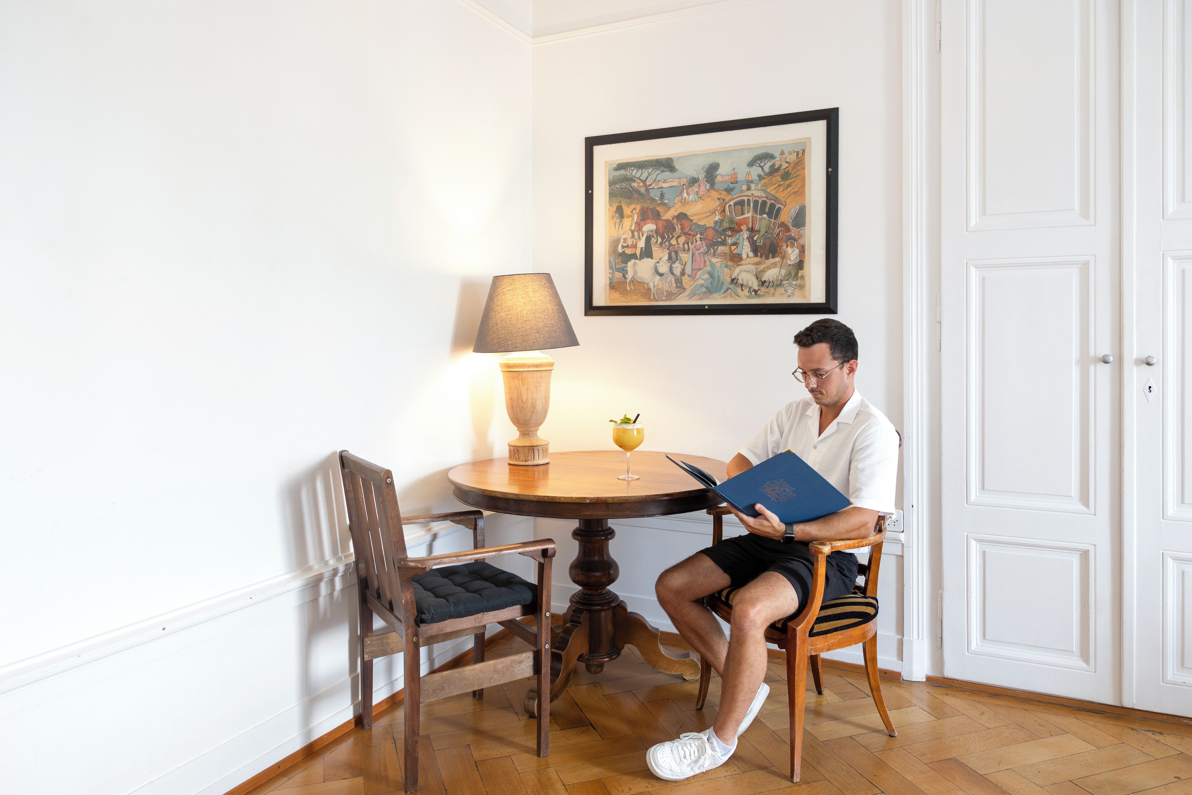 Un hombre leyendo un libro en una mesa de madera con una lámpara y un cuadro en la pared.