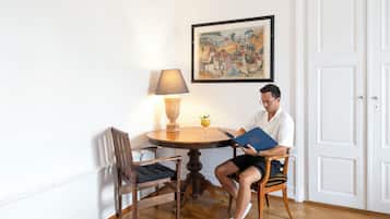 Un homme est assis à une table en bois, un livre à la main, une lampe sur la table et un tableau accroché au mur.