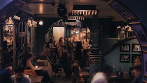 A bar area with people sitting and standing, wooden furniture, and a staircase.