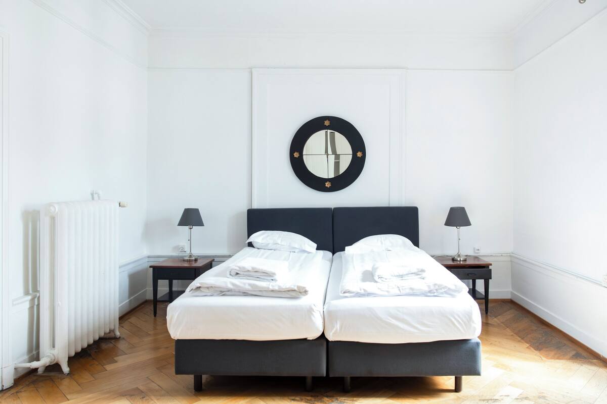 Una cama doble con ropa blanca, dos mesitas de noche con lámparas, un espejo en la pared y un radiador.