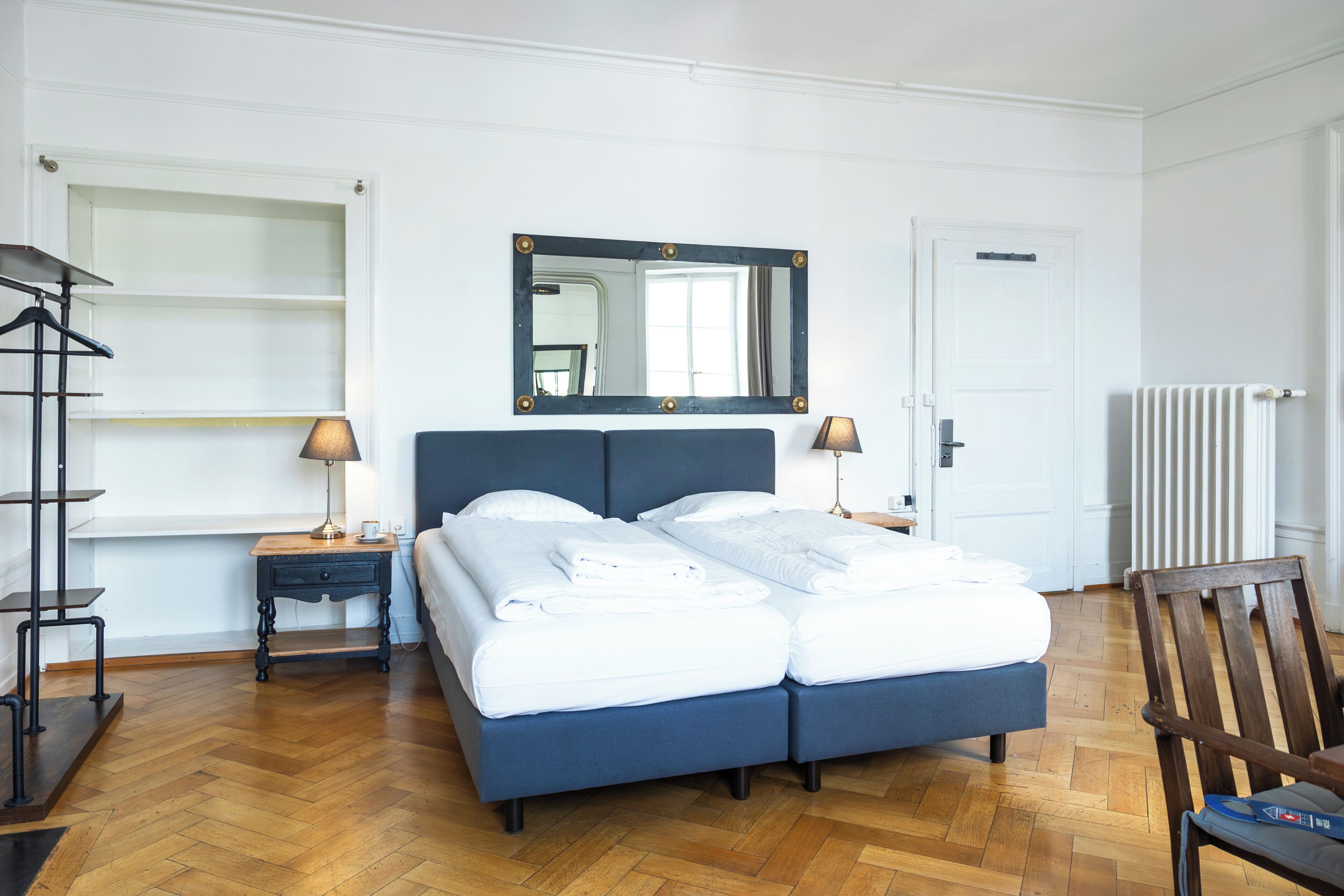 Un dormitorio ordenado con una cama doble, mesitas de noche con lámparas, un espejo y un radiador.