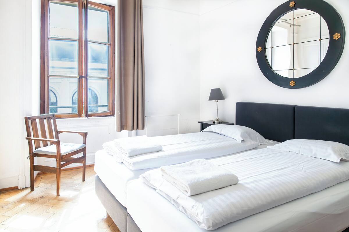 Habitación de hotel con dos camas, una silla, un espejo redondo y una ventana con cortinas.