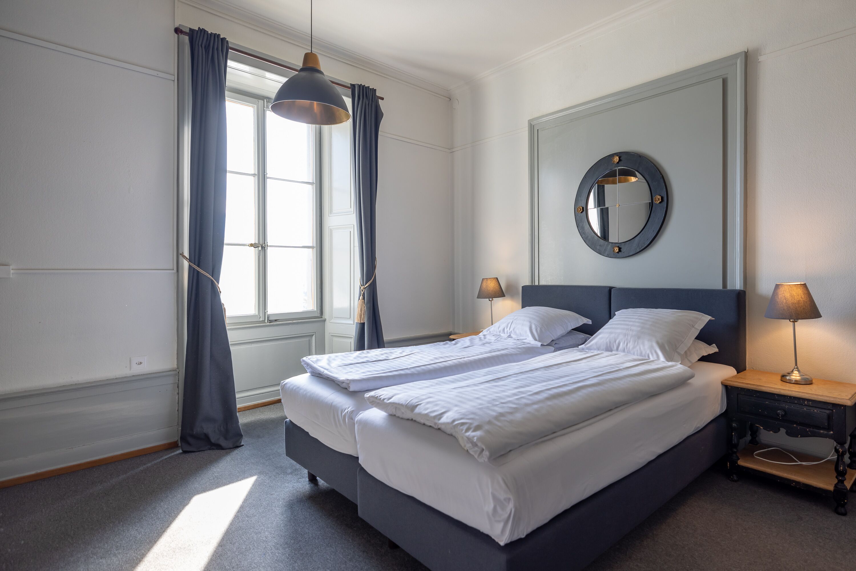 Ein Doppelbett mit weißer Bettwäsche, ein runder Spiegel an der Wand und zwei Nachttischlampen.