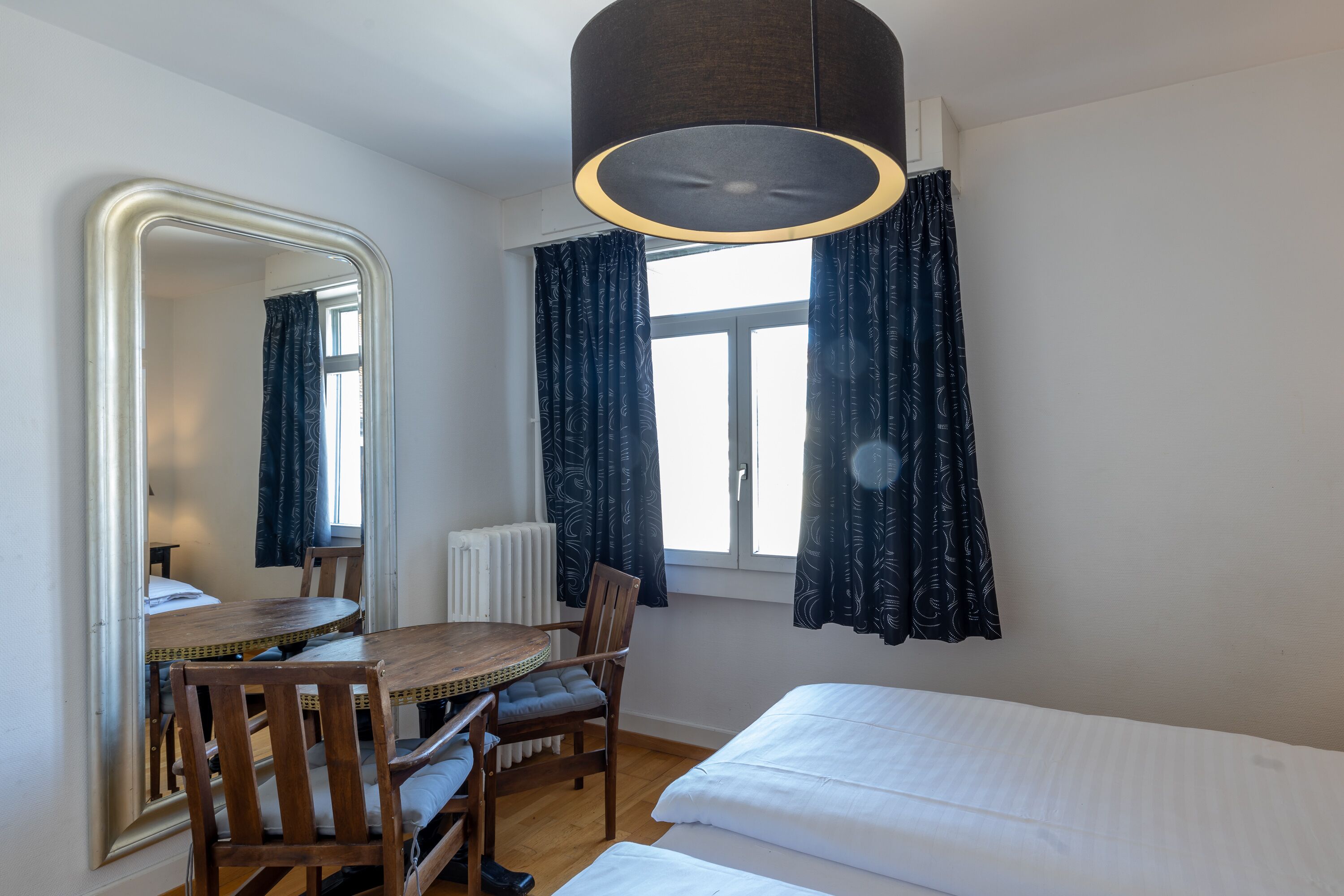 A room with a bed, a wooden table with chairs, a mirror, a window with curtains, and a ceiling light fixture.