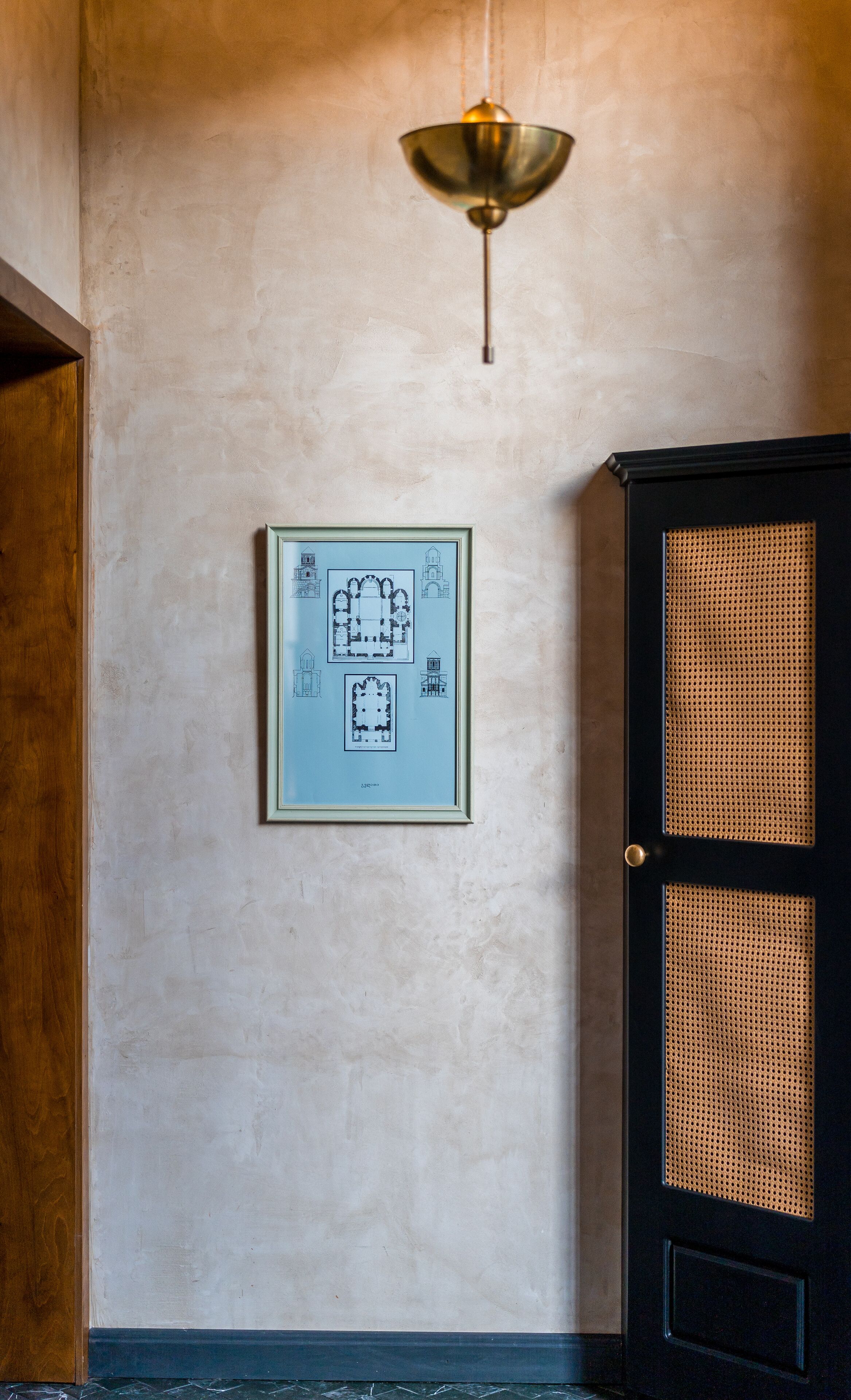 A framed architectural drawing on a textured wall, a hanging light fixture, and a black cabinet with a woven front panel.