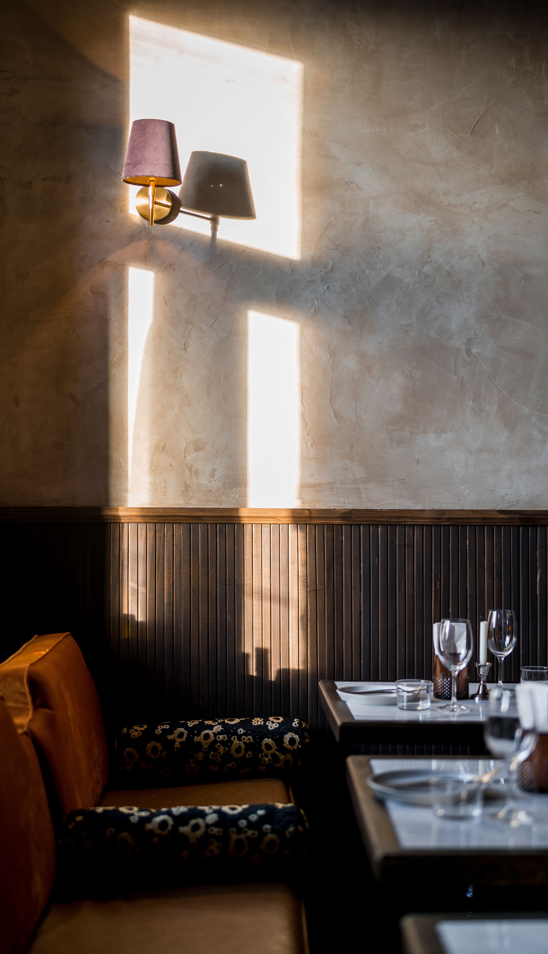 Una habitación tenuemente iluminada con una mesa, un banco, una lámpara de pared y una ventana que proyecta luz sobre la pared.