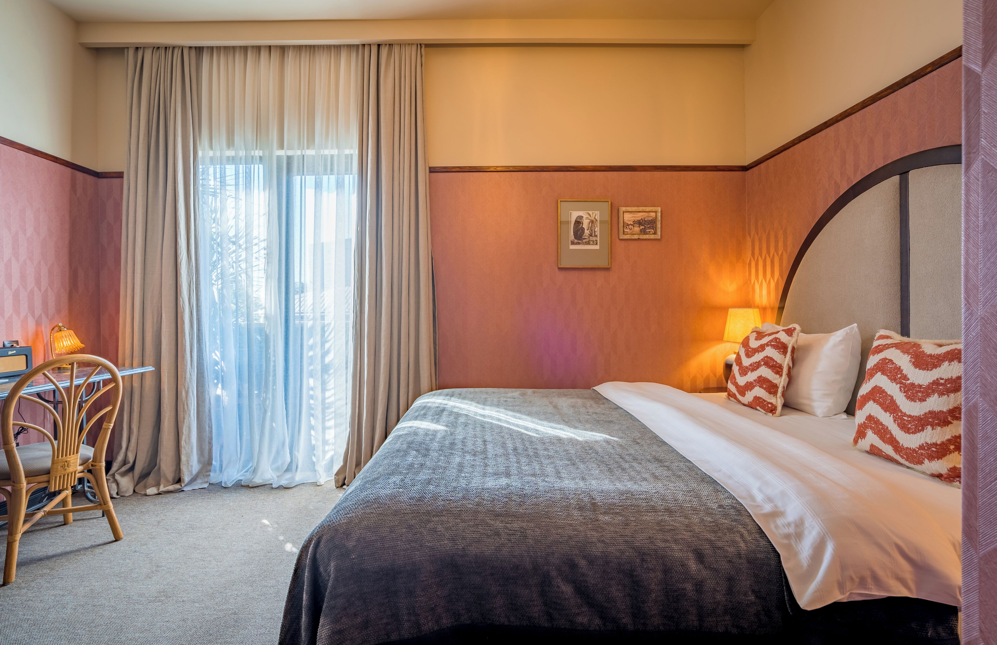 A hotel room with a bed, a desk, and a chair. There is a window with curtains, a lamp, and a framed picture on the wall.