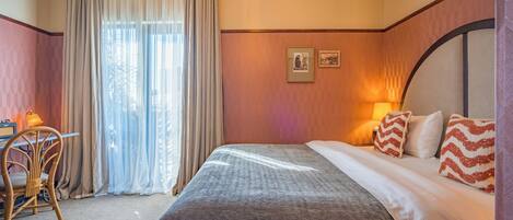 Habitación de hotel con cama, escritorio y silla. Hay una ventana con cortinas, una lámpara y un cuadro en la pared.