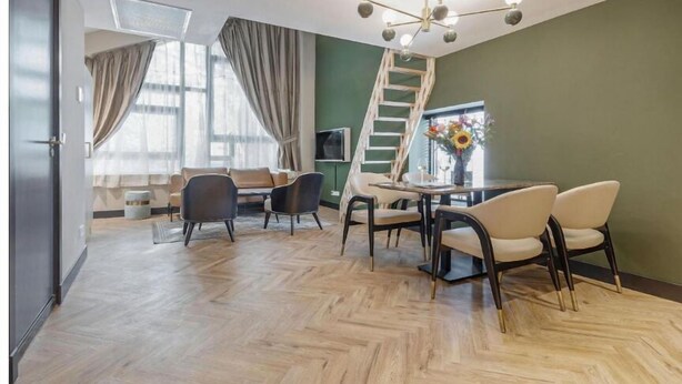 A modern living room with a herringbone floor, a dining area with a round table and chairs, a staircase, and a large window with curtains. - Bens The Walton (Amsterdam)