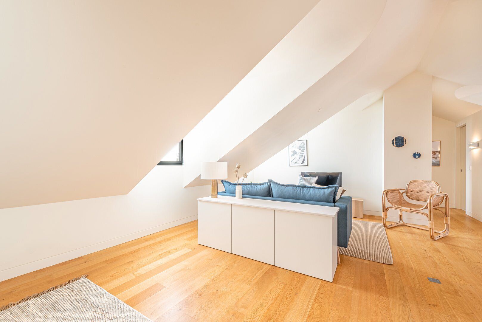 Un dormitorio tipo loft moderno con techo inclinado, una cama blanca con ropa de cama azul, una silla de mimbre y un aparador blanco.