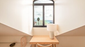 A room with a wooden desk, a lamp, a window with a view of the beach, and a small wicker basket. - GuestReady - Conde das Acáçovas 3 (Paço de Arcos)