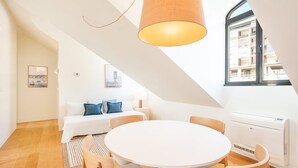A modern, well-lit room with a white round table and chairs, a bed with blue pillows, and a large window offering a view of buildings. - GuestReady - Conde das Acáçovas 3 (Paço de Arcos)