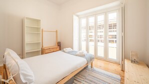 A bedroom with a bed, a wooden wardrobe, a small table, and a window with a view of buildings.