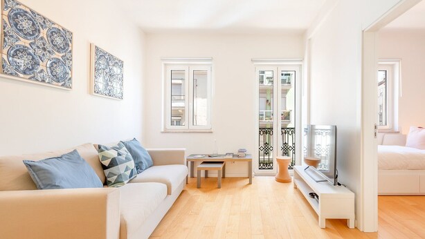 Ein modernes Wohnzimmer mit einer Couch, einem Couchtisch und einem Fernseher. Man sieht zwei Fenster und einen Balkon.