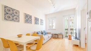 Ein helles Wohnzimmer mit einem weißen Sofa, gelben Sesseln, einem Esstisch und einem Fernseher auf einem Schrank.