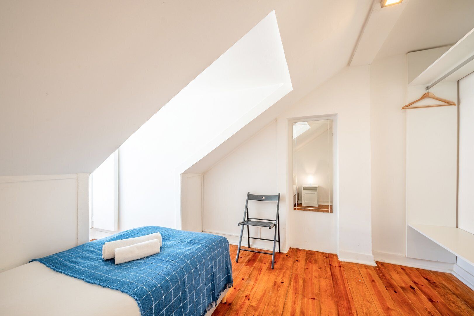 A bedroom with a sloped ceiling, a bed with a blue blanket, a chair, and a mirror on the wall.