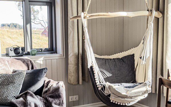 A cozy room with a hanging hammock chair, a window with a view of a field, and a radio on the windowsill. - Askot Cottages (Ásahreppur)
