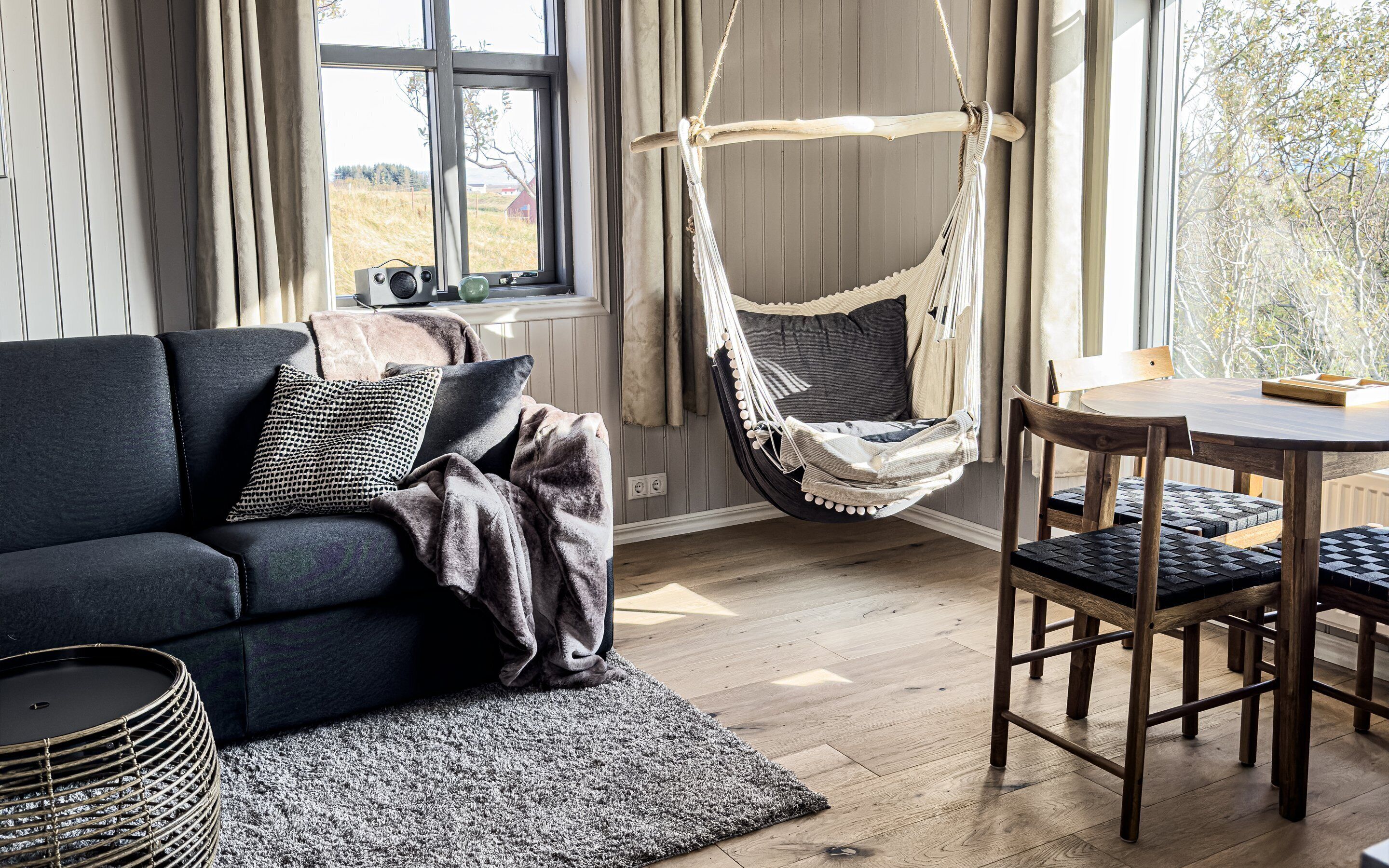 A cozy living room with a dark sofa, a hanging chair, a wooden dining table, and a view of the outdoors through large windows.