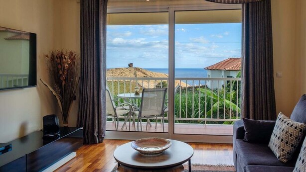 A room with a large sliding glass door leading to a balcony with a view of the sea and greenery.