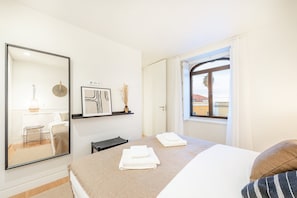 A hotel room with a bed, a bedside table, a mirror, a window with a view of buildings, and a shelf with decorative items. - GuestReady - Conde das Acáçovas 1E (Oeiras)