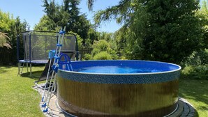 A circular swimming pool with a wooden exterior and a blue interior, surrounded by a brick border, in a backyard with a trampoline and a ladder. - Residence Dymokury (Stredoceský kraj)