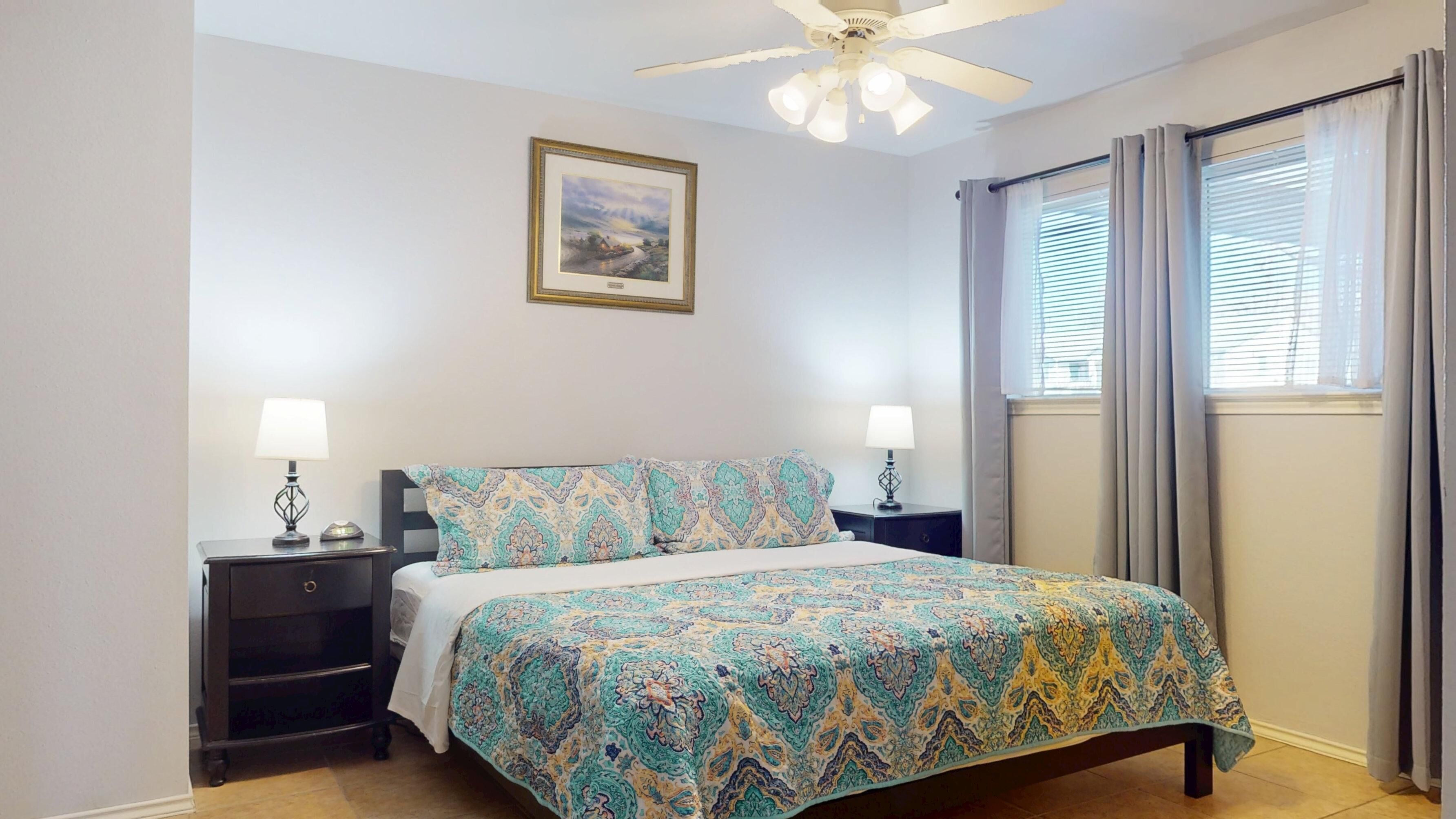 Une chambre à coucher comprenant un lit, deux tables de chevet, un ventilateur de plafond, une fenêtre avec des rideaux et un tableau encadré accroché au mur.