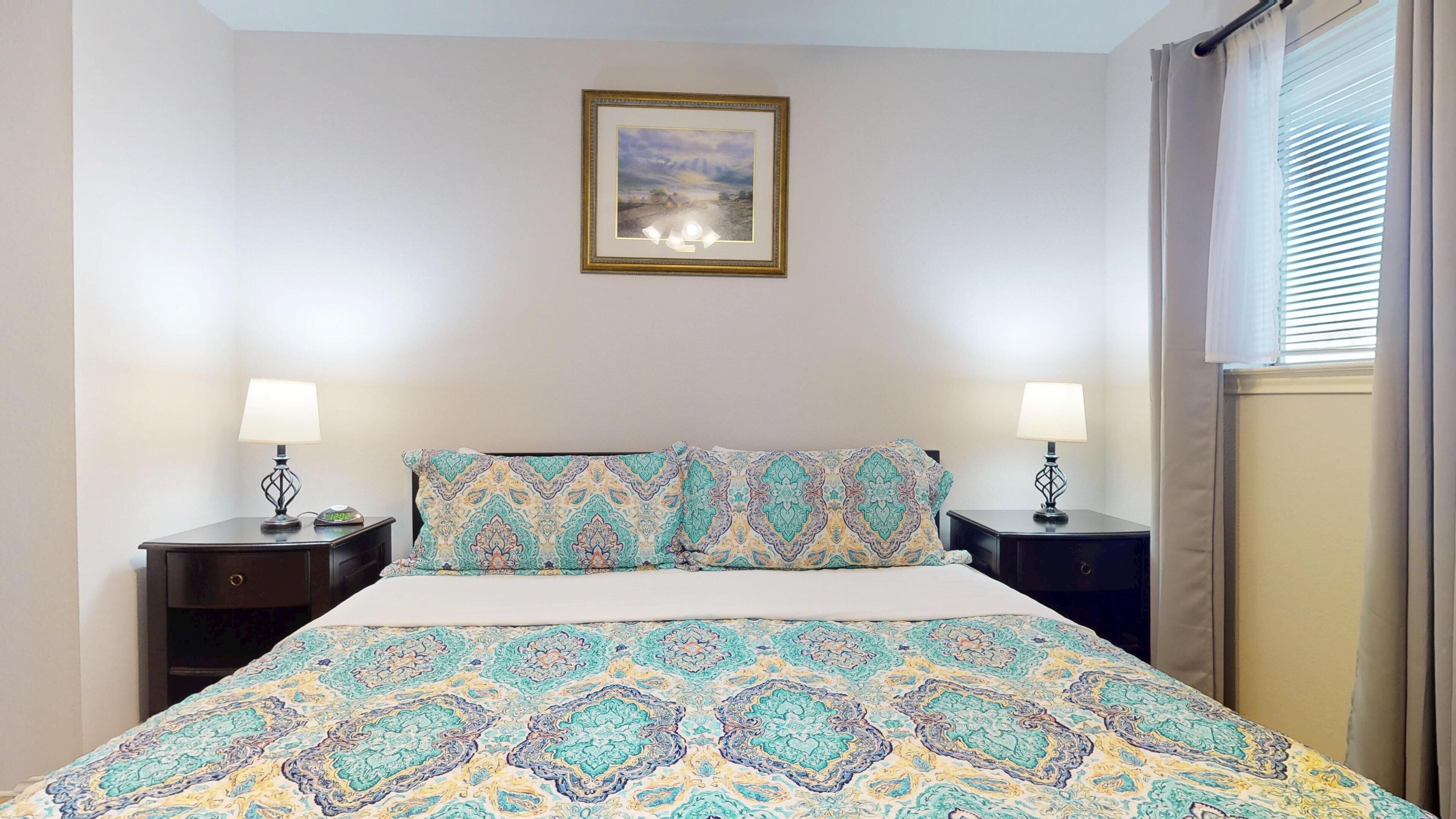 Une chambre à coucher avec un lit, deux tables de chevet, une commode, un tableau encadré au mur et une fenêtre avec des rideaux.